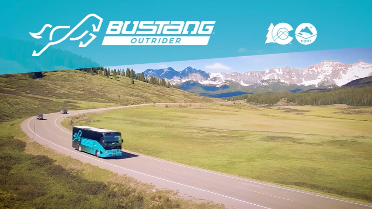 A Bustang Outrider coach on a highway through green ranchland, with mountains behind.