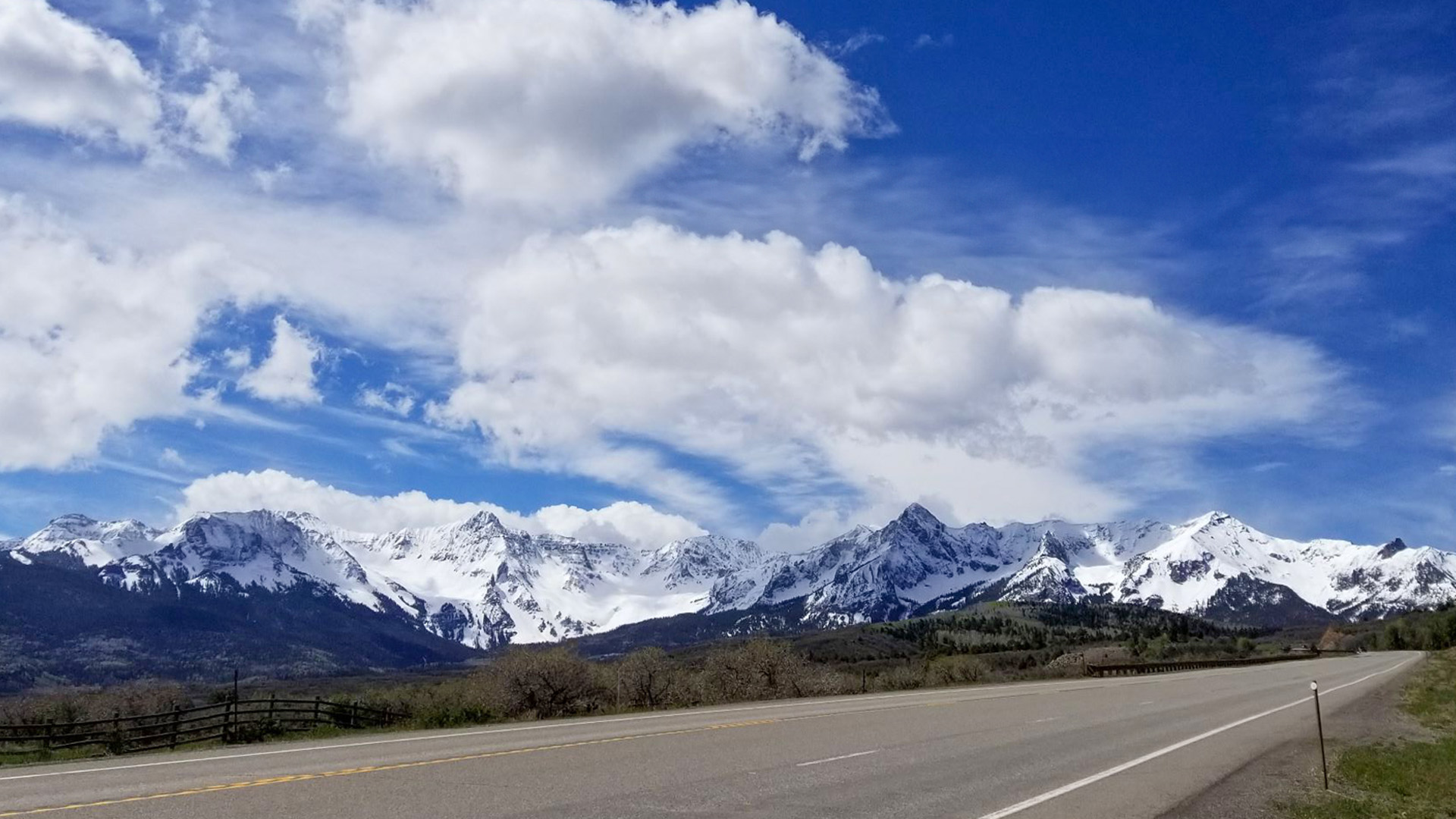 A two-lane highway curving toward snow-capped Colorado mountains under a blue sky.