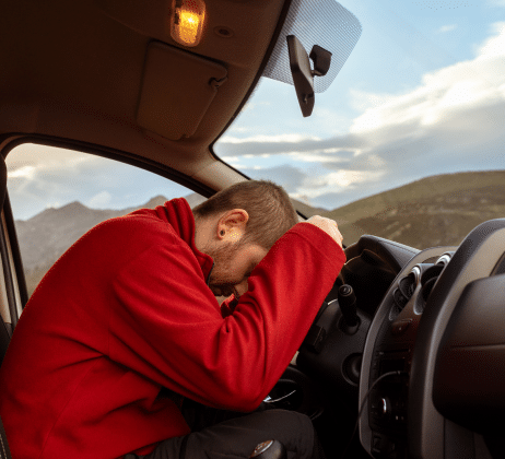 A man in a red hooded sweatshirt slumped forward against the steering wheel of a car.